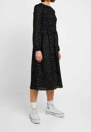 Osoba nosząca czarną sukienkę midi w białe groszki, z długimi rękawami oraz białe wysokie trampki z białymi skarpetkami.