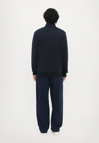 Sudadera navy con cuello alto, puños y dobladillo acanalados; combinada con pantalones de mezclilla oscuros de corte holgado y zapatillas crema.