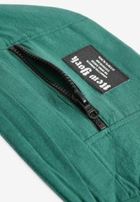 Groene stof met een gestructureerd oppervlak, voorzien van een zwarte rits en een zwart label met witte tekst, bevestigd aan het bovenste gedeelte.