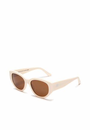 Gafas de sol con forma de ojos de gato en color crema con lentes marrones y un pequeño emblema plateado en la patilla, mostradas sobre un fondo blanco.