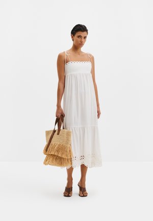 Femme portant une robe midi blanche sans manches avec des détails en œillets, tenant un sac fourre-tout tissé à franges, et chaussée de talons ouverts marron.
