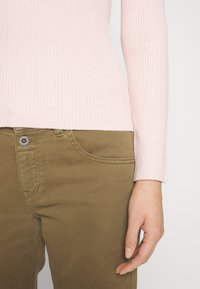 Donna che indossa un top a righe lunghe di un rosa chiaro e pantaloni verde oliva, mostrando il busto e la mano destra rilassata al lato.