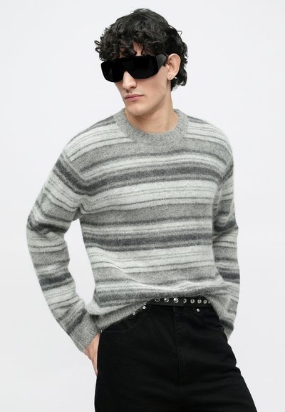 Sivi i bijeli džemper s prugama, okruglog izreza i dugim rukavima, uparen s crnim hlačama koje imaju remen sa zakovicama. Crne sunčane naočale.