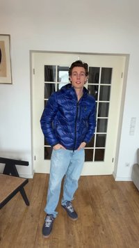 Jeune homme portant une doudoune bleue, un jean bleu clair et des baskets grises, debout à l'intérieur sur un sol en bois près de portes doubles blanches.