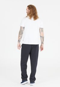 Vit t-shirt med korta ärmar, kombinerad med mörkgrå sweatpants, bärs av en person med tatueringar, sedd bakifrån.