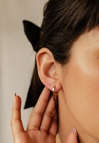 Złoty kolczyk w kształcie koła na przekłutej uchu, o polerowanym wykończeniu. W tle widać ciemne włosy, częściowo związane czarną wstążką.