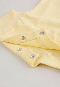 Prenda amarilla para bebé con tres botones a presión plateados parcialmente abiertos sobre una superficie lisa.