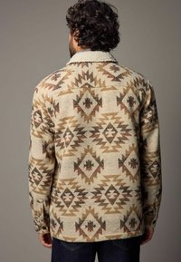 Veste chemise beige avec un design texturé et imprimé présentant des formes géométriques en marron et noir. Fermée par un bouton au niveau du poignet.