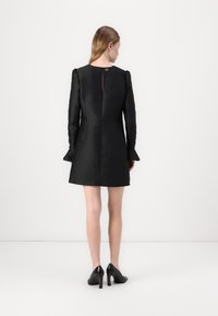 Mini abito nero con texture, maniche lunghe, con uno spacco profondo sul retro e polsini svasati, abbinato a scarpe con tacco alto nere.