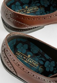 Pantof brown din piele, cu detalii perforate, și o branț cu model floral albastru, având text de branding auriu, arătând vârful rotunjit și accente cusute.