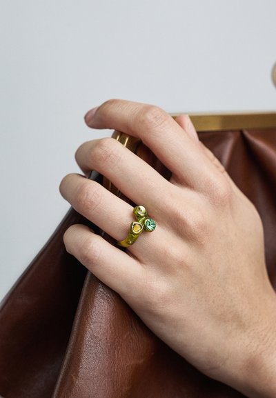 Roka tur brūnu ādas maku, nēsājot zelta gredzenu ar zaļiem un dzeltens dārgakmeņiem uz gredzena pirksta.