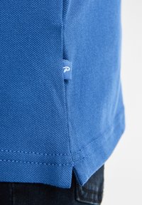 Modrá bavlněná košile se strukturovaným povrchem, s malou značkovou etiketou a bočním rozparkem pro snadnější pohyb.