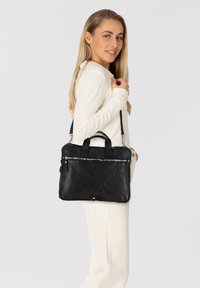 Bolso de cuero negro con dos asas y una correa ajustable, que cuenta con un cierre de cremallera y una superficie texturizada. Diseño rectangular sencillo.