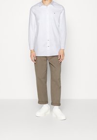 Camisa blanca de botones con mangas largas y un pequeño logo en el pecho, combinada con chinos color marrón claro arremangados y zapatillas blancas.