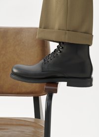 Botín negro de cuero hasta el tobillo con puntera redondeada, superficie texturizada, diseño de cordones y suela negra gruesa, descansando sobre una silla de madera.