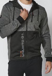 Sweat à capuche zippé avec un mélange de tissu gris foncé et noir. Présente une partie supérieure texturée, une partie inférieure noire unie, et un détail de logo orange.
