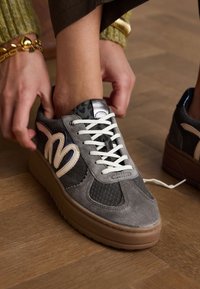 Gray suede sneakers with textured black accents, white laces, and a round toe. Features a smooth brown sole and metallic detailing.