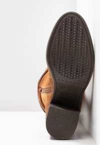 Brown suede boot displayed from the bottom, featuring a textured black rubber sole with a woven pattern and a size label.