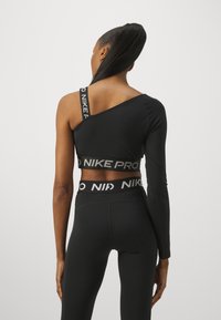 Asymmetrisk svart sportsoverdel med én lang erm, hvit logo-bånd, og høyt midjet svarte tights med kontrasterende logo på midjebåndet.