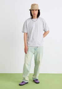 T-shirt cinza de algodão, jeans baggy azul claro, tênis roxos e um chapéu de balde bege. Colar de contas, em pé num chão verde.