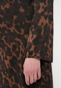 Material maro cu un model de imprimeu leopard, cu pete negre pe un fundal maro deschis. Mâneci lungi cu croială lejeră.