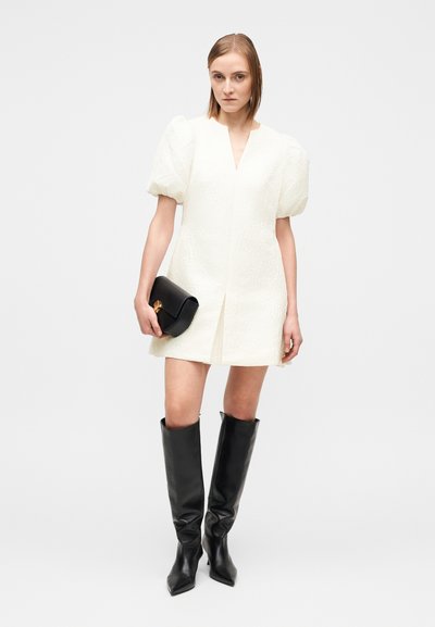 Vestido branco texturizado com mangas bufantes e decote em V. Combinado com uma clutch preta e botas pretas de pele até ao joelho.