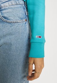 Ljusblå ribbad tröjärm med varumärkesmärke, i kombination med ljus tvättade denimjeans. Guldringar pryder handen.