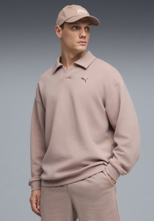 Hombre con gorra Puma beige, polo de manga larga beige y pantalones a juego, mirando ligeramente hacia la derecha con una mano en el bolsillo.