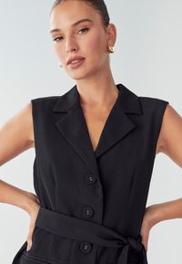 Czarna sukienka bez rękawów z kołnierzykiem, przednimi guzikami oraz związanym detalem w talii, wykonana z gładkiego materiału o matowym wykończeniu.