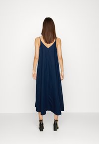 Mujer con cabello castaño liso vestida con un vestido midi sin mangas de color azul marino y sandalias de tacón negro, de pie contra un fondo blanco liso.