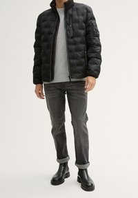 Černá péřová bunda s prošívaným designem, předním zipem a vysokým límcem, přes šedý svetr, kombinovaná s tmavými džíny s vyhrnutými lemy a černými kotníkovými botami.