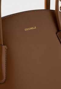 Bolso de mano de cuero marrón con textura suave, que presenta un logo dorado "Coccinelle". El diseño incluye lados estructurados y asas resistentes.