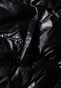 Panel de chaqueta tipo plumón negra con textura brillante y acolchada, con costuras visibles y un bolsillo con cremallera lateral. Material suave y reflectante.