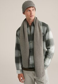 Beanie gri imună cu dungi și fular asortat purtate cu o cămașă flanelată verde cu carouri, cu accente negre și albe, și pantaloni gri deschis.