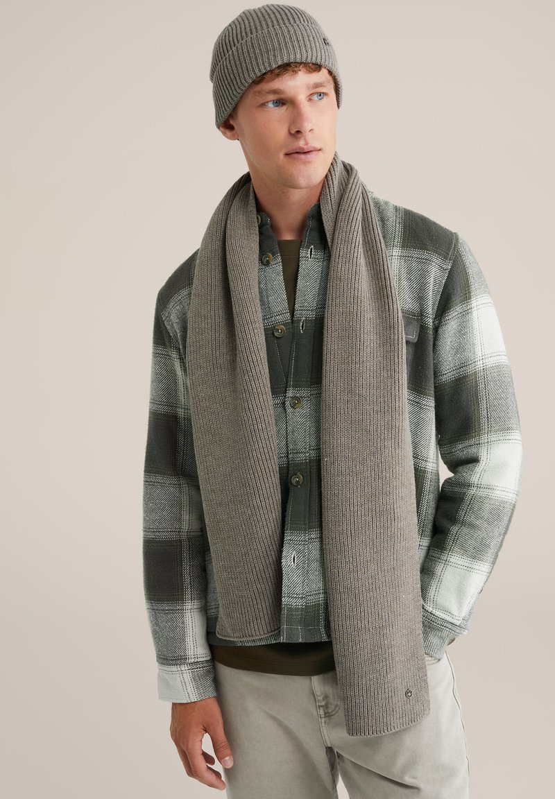 Beanie gri imună cu dungi și fular asortat purtate cu o cămașă flanelată verde cu carouri, cu accente negre și albe, și pantaloni gri deschis.