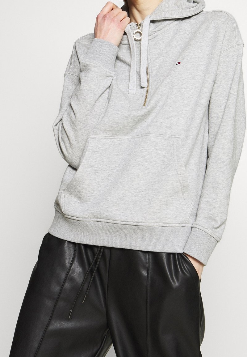 Sweat à capuche gris en matière douce avec une poche avant, une capuche à cordon de serrage et un détail zippé. Assorti à un pantalon en cuir noir.