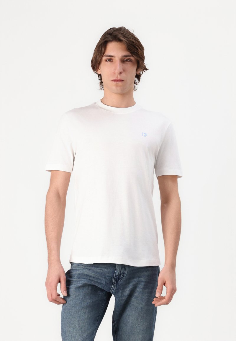 Camiseta blanca de manga corta con escote redondo y un pequeño logotipo azul claro en el lado izquierdo del pecho. Tejido de algodón suave. Llevada con jeans azules.