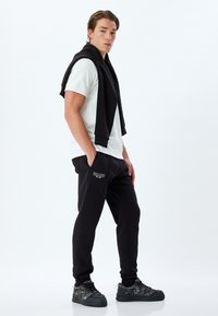 Svarta joggers med en vit logotyp, kombinerat med en vit t-shirt och en svart sweatshirt över axlarna, stylat med svarta skor.