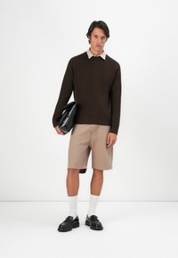 Brauner Strickpullover, hellbeige Hemd mit Kragen, beigefarbene, gefaltete Shorts und schwarze Loafer. Das Model hält eine schwarze Clutch, steht vor einem schlichten Hintergrund.