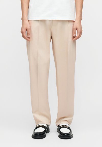 Pantalon beige clair sur mesure avec une coupe ample, présentant une texture lisse, des plis simples et une coupe droite. Porté avec des mocassins noirs.