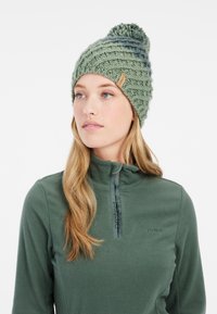 Cappello in maglia verde con un motivo testurizzato e un pompon in cima. Presenta un'etichetta di tessuto sul lato. Indossato con una giacca in fleece verde.