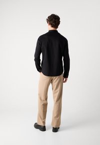 Zwarte button-up shirt met lange mouwen, rechte snit en kraag, gecombineerd met beige broek en zwarte schoenen, gezien vanuit de achterkant.