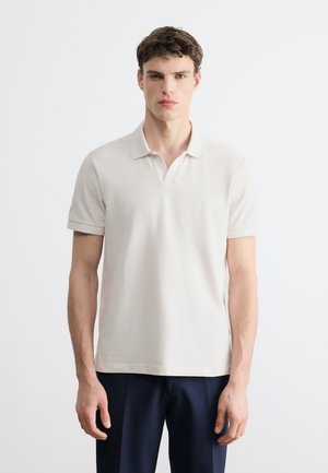 Giovane uomo con capelli ricci e scuri, indossa una polo a maniche corte color avorio e pantaloni scuri, in piedi contro uno sfondo bianco semplice.