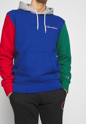 Man draagt een blauwe Champion-trui met een rode linker mouw, groene rechter mouw, grijze capuchon met witte trekkoorden en een zwarte broek.