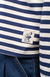 Niebiesko-biała w paski bawełniana koszulka z metką zawierającą wzór postaci i tekst "KAOTIKO SOCIETY" naszyty na dolnym brzegu.