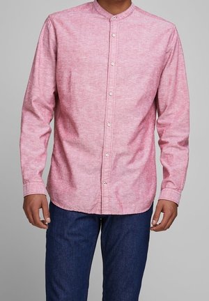 Homme portant une chemise rose à manches longues avec col mandarin et un jean bleu foncé, debout devant un fond gris.
