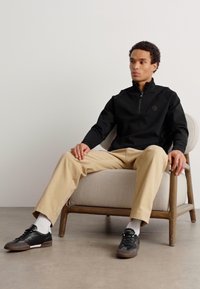 Felpa nera con zip e logo, pantaloni beige e sneakers neri. Seduto su una sedia di legno con un cuscino chiaro.