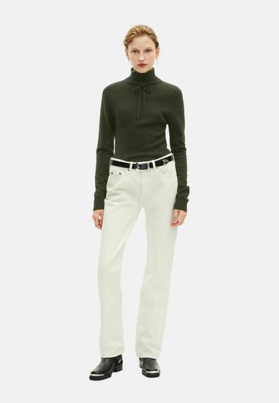 Suéter de cuello alto verde oscuro con detalle de cordón, combinado con pantalones rectos en un tono blanco roto y botines negros con acentos metálicos.