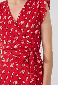 Red wrap dress with a textured fabric, featuring a floral pattern in shades of white and black. Ruffled sleeves and tied waist detail.