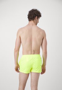 Pantaloni de baie neon galbeni, cu talie elastică și un buzunar la spate. Țesătura pare ușoară și are o textură netedă.